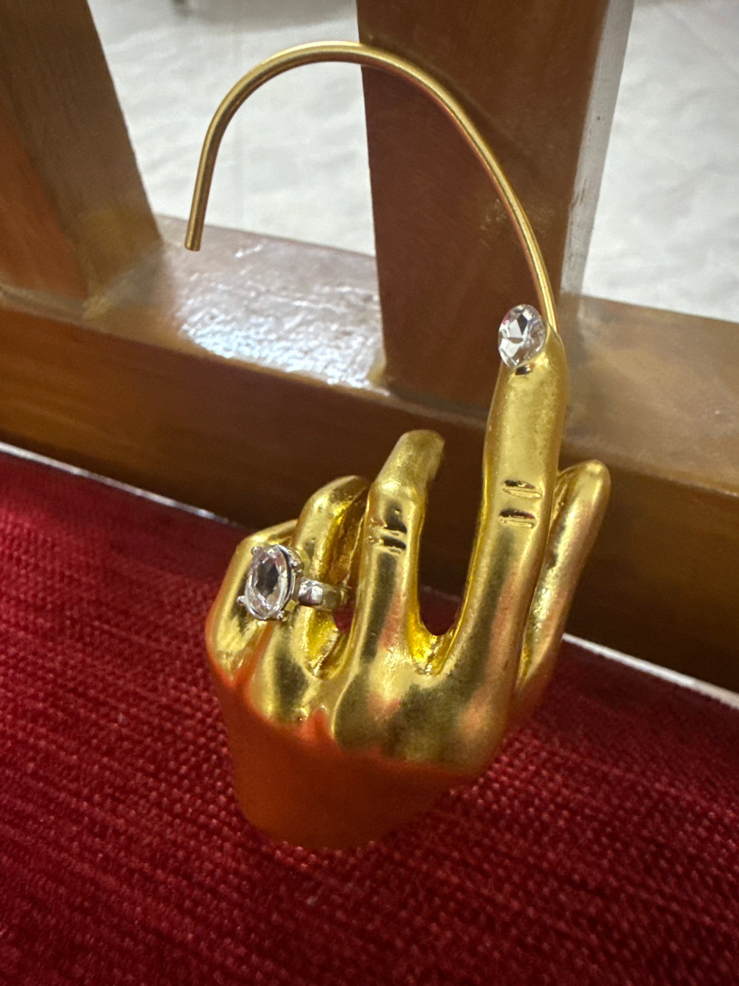 A gold-tone hand-shaped jewelry display stand holding two clear gemstone rings, photographed on a red velvet surface with a wooden chair back in the background.