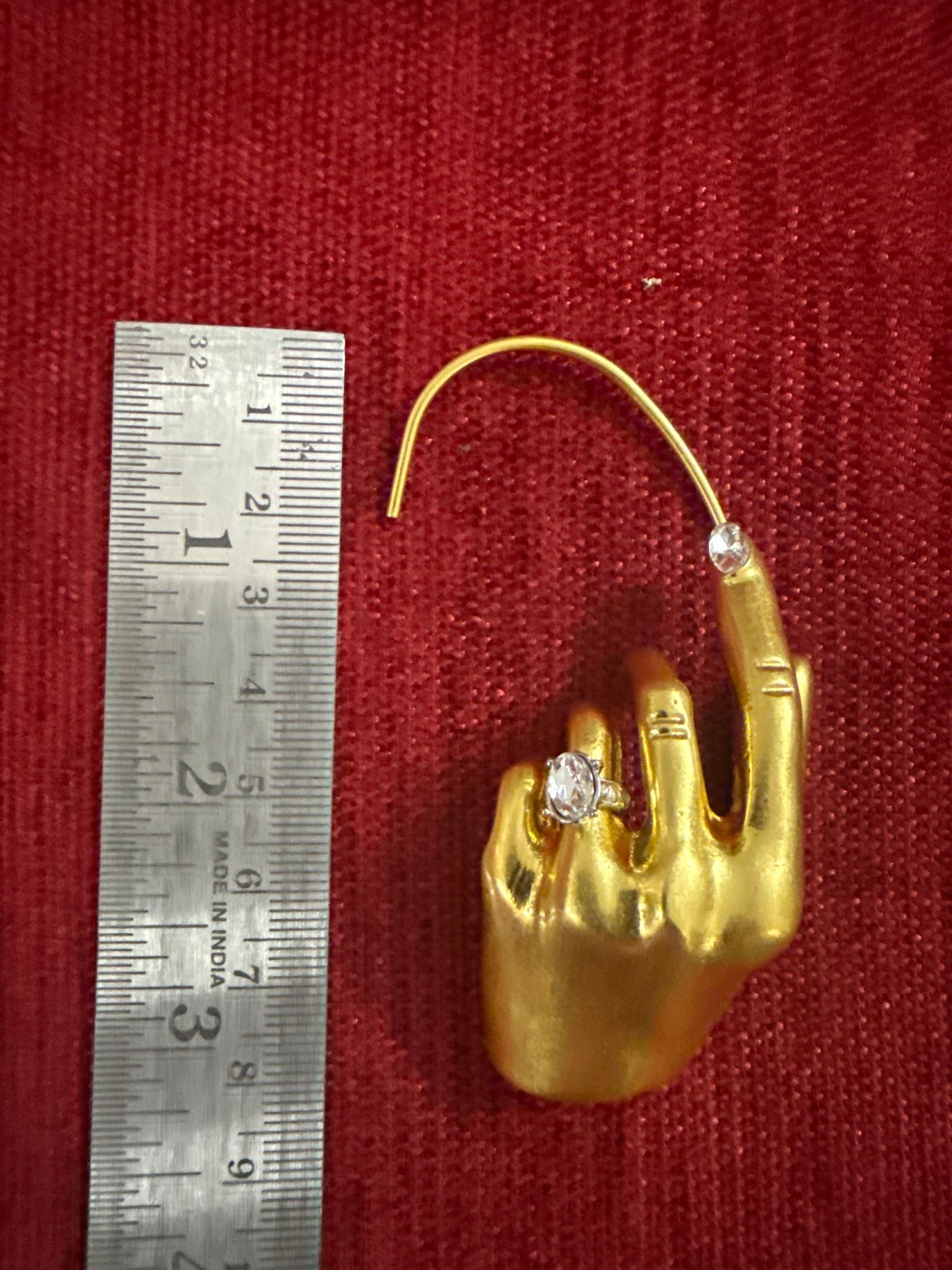 A gold hand-shaped ring holder or jewelry display piece with a curved loop on the index finger, shown next to a metal ruler on a red textured background. The piece resembles a hand wearing a ring, with clear gemstones on the fingers.