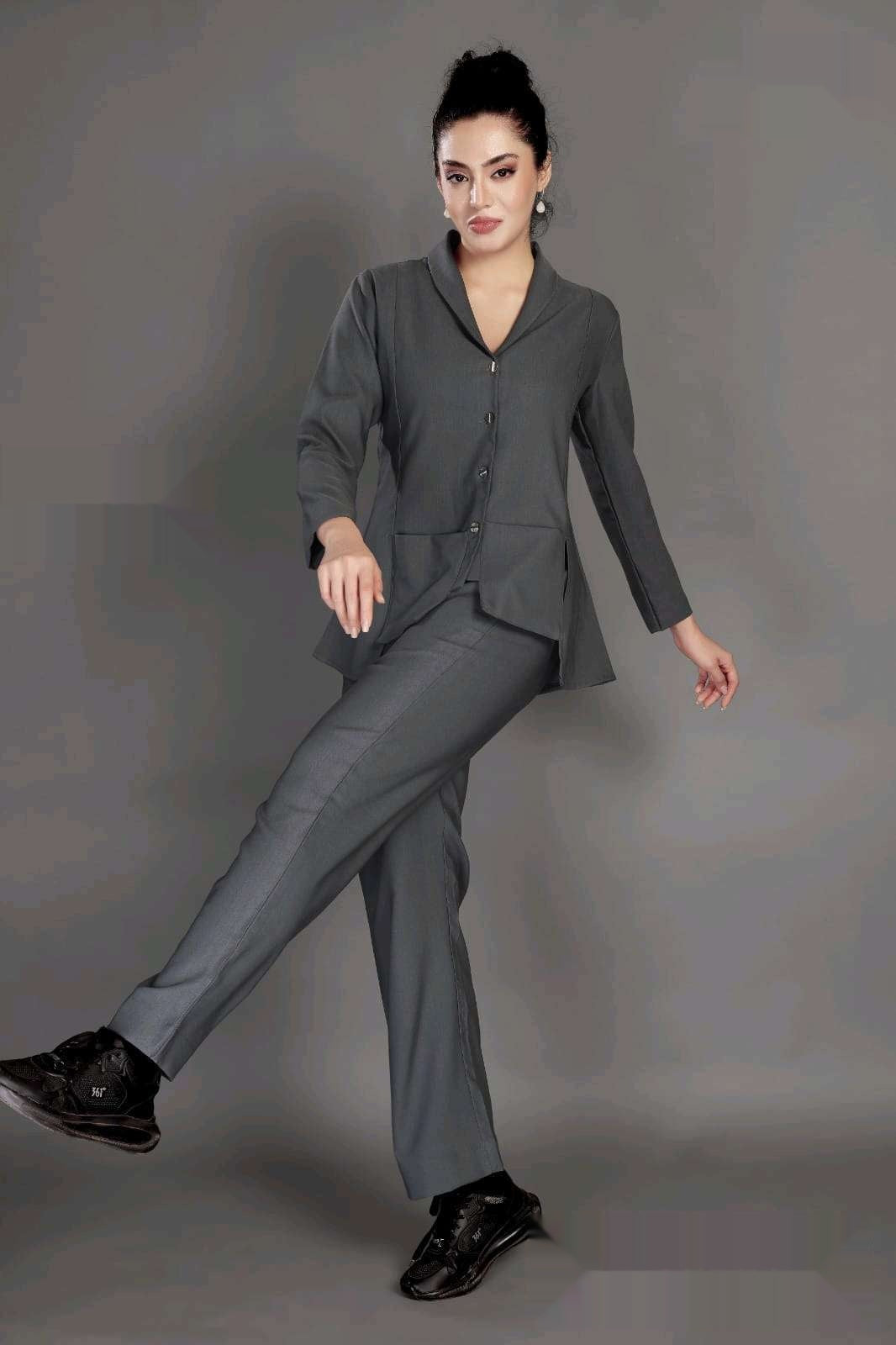 A woman modeling a dark gray two-piece suit consisting of a long-sleeve button-front blazer and matching trousers, paired with black sneakers, against a solid gray studio background.