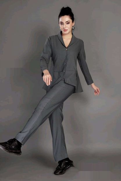 A woman modeling a dark gray two-piece suit consisting of a long-sleeve button-front blazer and matching trousers, paired with black sneakers, against a solid gray studio background.