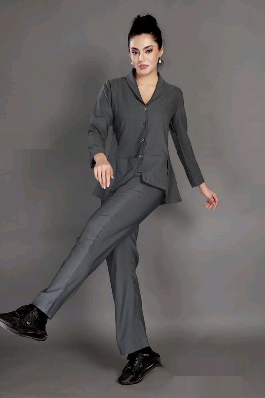 A woman modeling a dark gray two-piece suit consisting of a long-sleeve button-front blazer and matching trousers, paired with black sneakers, against a solid gray studio background.
